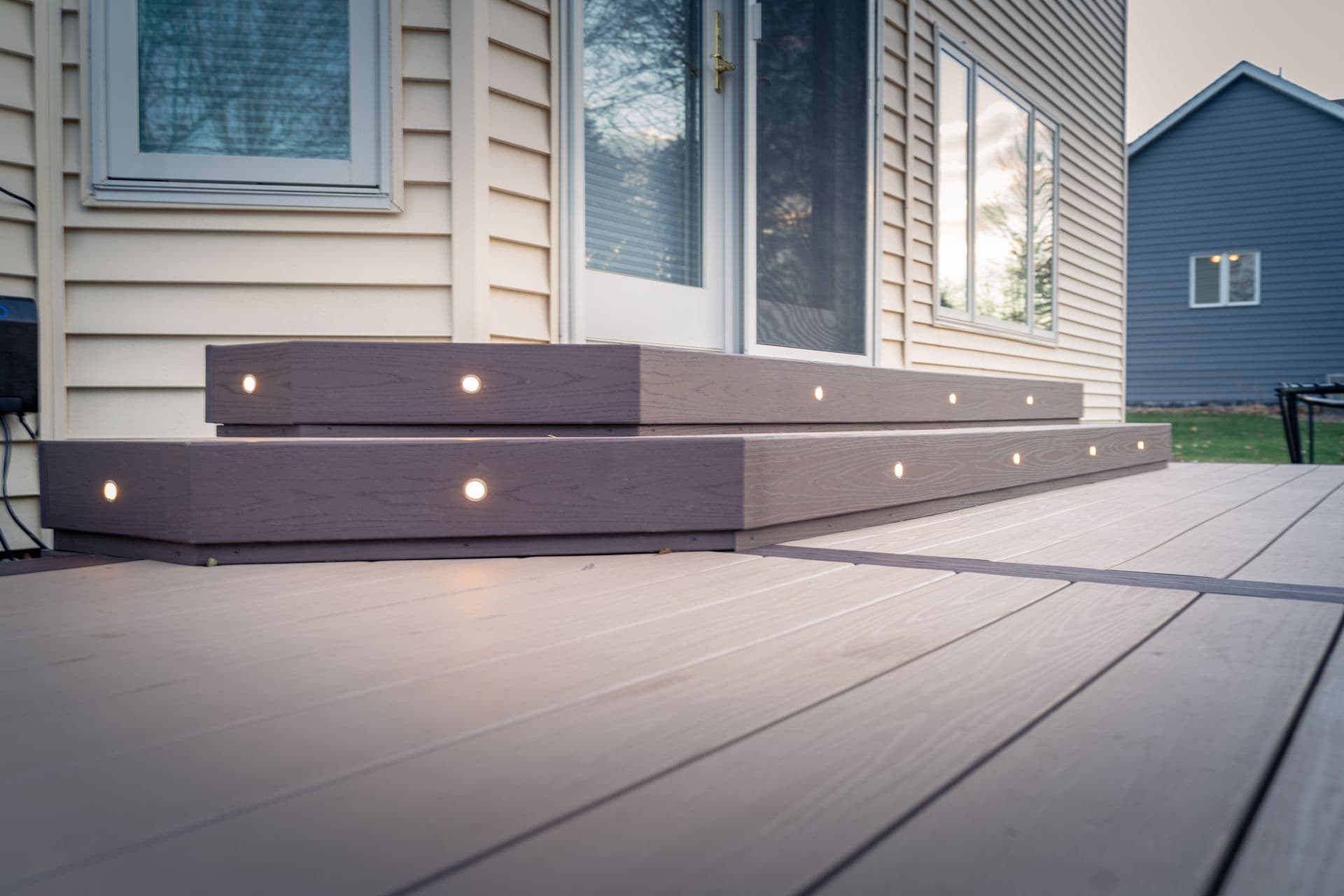Low-angle view of two wide composite deck steps outside a cream-colored house, featuring dark risers with circular recessed LED lights built into them.