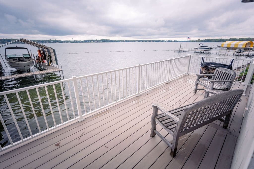 View of a lakeside composite deck with white railings and weathered wooden benches overlooking a boat dock under an overcast sky.