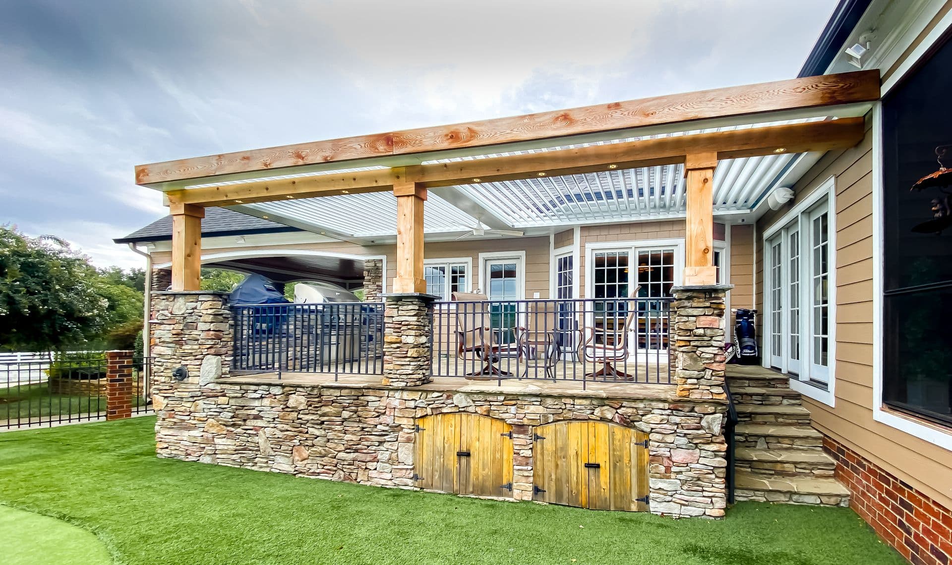 Cedar-Wrapped Louvered Pergola on Stone Patio Base (cedar wrapped pergola-3.jpg) A wide side perspective view of an elevated stone-veneer patio with built-in wooden storage doors underneath, supported by stone pillars topped with natural cedar wood-wrapped posts and a matching cedar beam frame enclosing a white louvered pergola roof system with integrated recessed lighting and a ceiling fan, attached to a tan siding home.