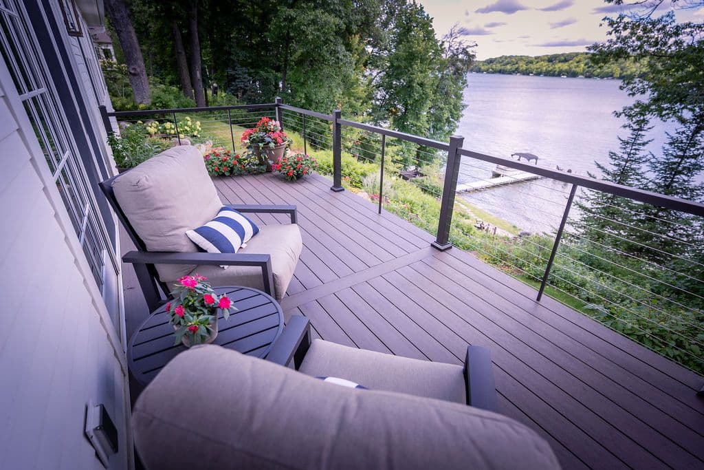 Elevated perspective view of a composite deck with cable railings, cushioned outdoor lounge chairs, side table, and potted flowers overlooking a scenic lake.