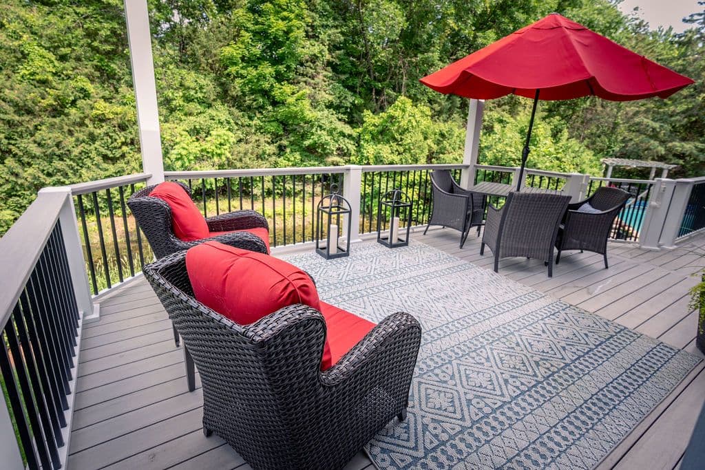 Elevated perspective of a grey composite deck with dark railings, a patterned outdoor area rug, dark wicker armchairs with bright red cushions, and a patio dining table with a red umbrella overlooking lush green trees.