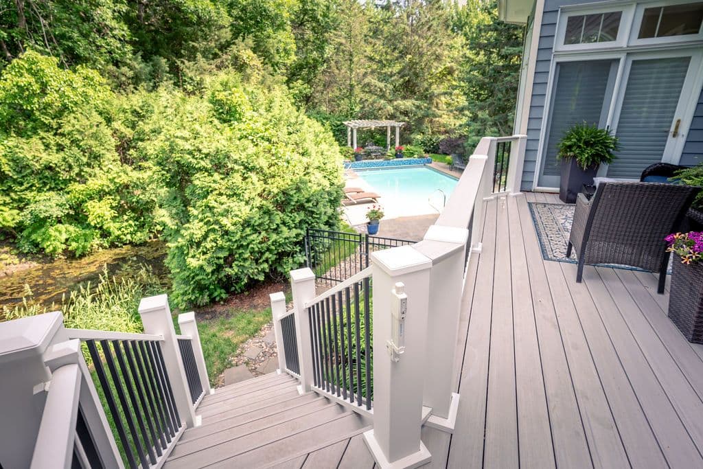 High-angle perspective of light composite deck stairs leading down toward a backyard swimming pool surrounded by greenery.
