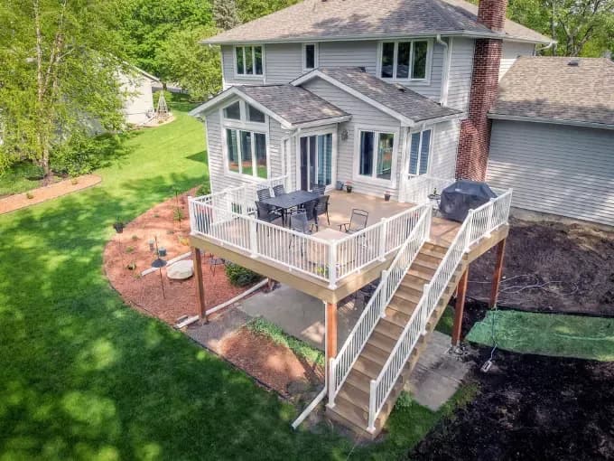 Elevated aerial view of the back of a two-story light grey siding home featuring a large raised composite deck with white railings, outdoor patio furniture, a grill, and wooden stairs leading down to a manicured lawn and garden beds.