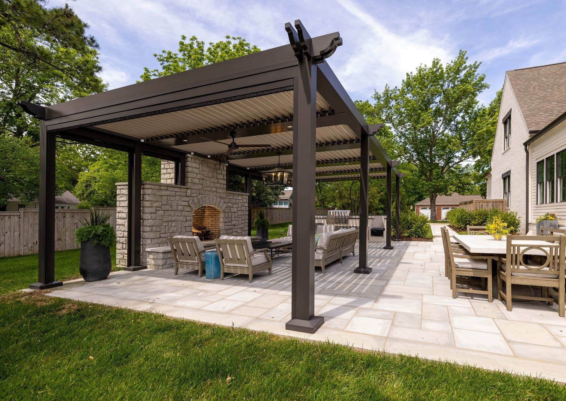 Low-angle perspective view of a spacious stone patio adjacent to a white house, covered by a black metal louvered pergola with decorative beam ends, featuring an integrated outdoor stone fireplace, ceiling fan, lounge seating, and an adjacent outdoor dining table.
