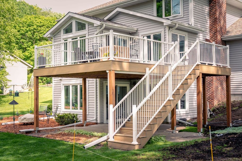 Elevated view of a two-story grey siding home featuring a second-story composite deck with white railings and staircase leading down to a lower patio area.