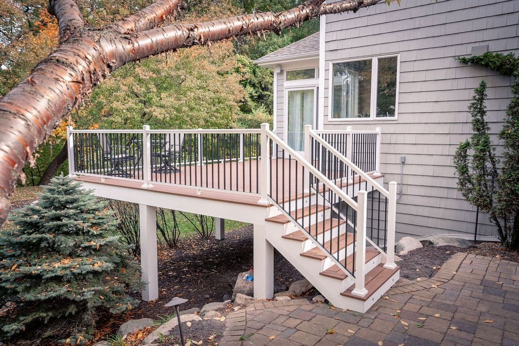 Angled view of an elevated composite deck attached to a shingled house, featuring white posts, black balusters, and stairs leading down to a paver patio.