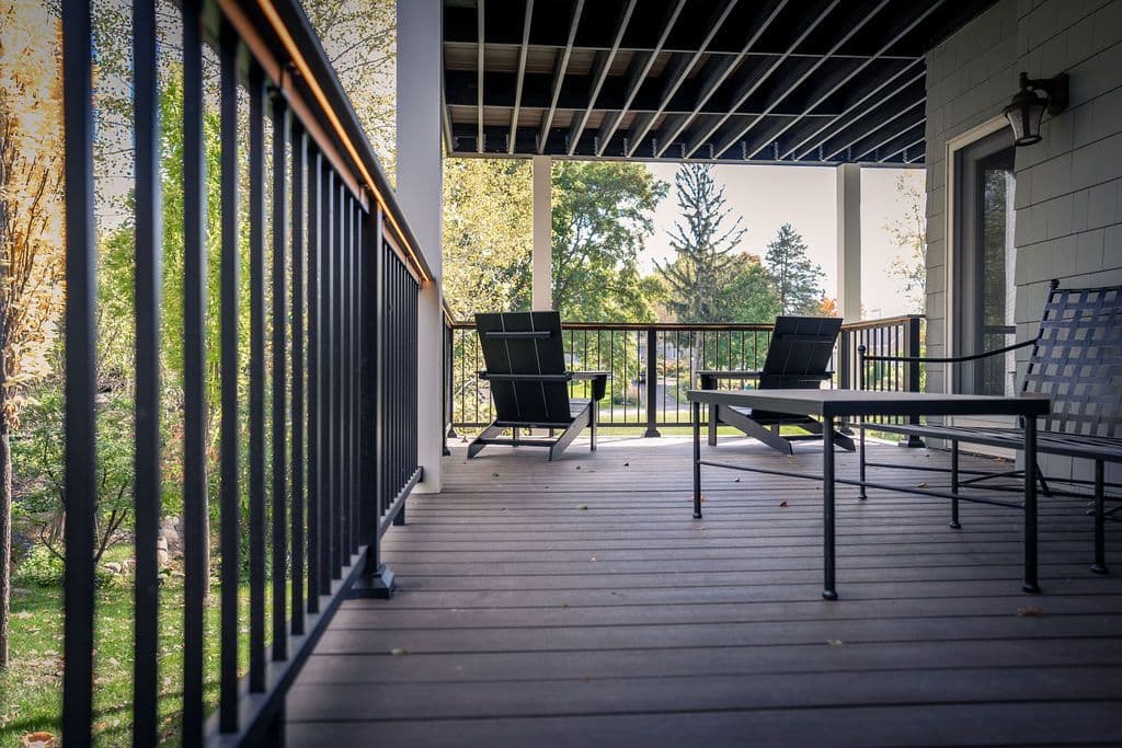 Eye-level perspective under a covered deck featuring composite flooring, dark metal railings, and black outdoor lounge chairs looking out toward a green yard.