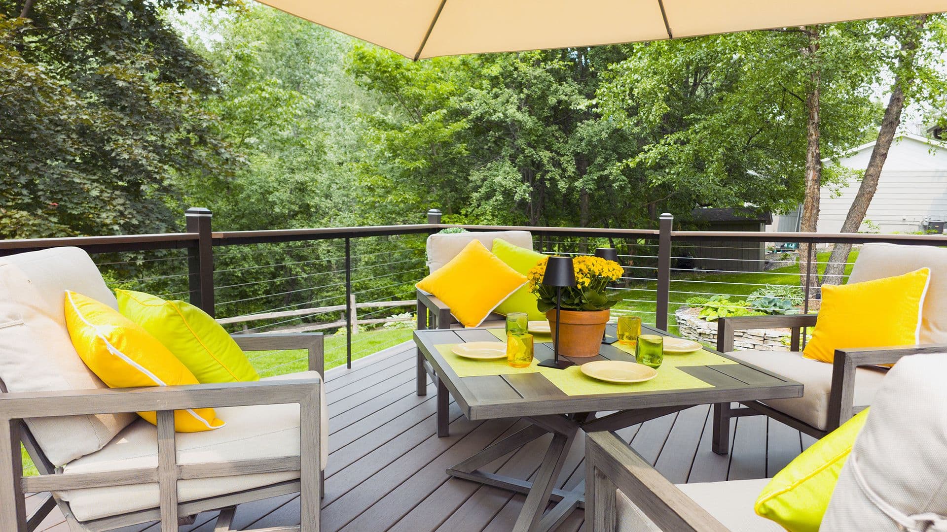 Eye-level view of a composite deck dining area with modern wooden armchairs, bright yellow and lime throw pillows, a potted flower centerpiece, and cable railings overlooking lush green trees.