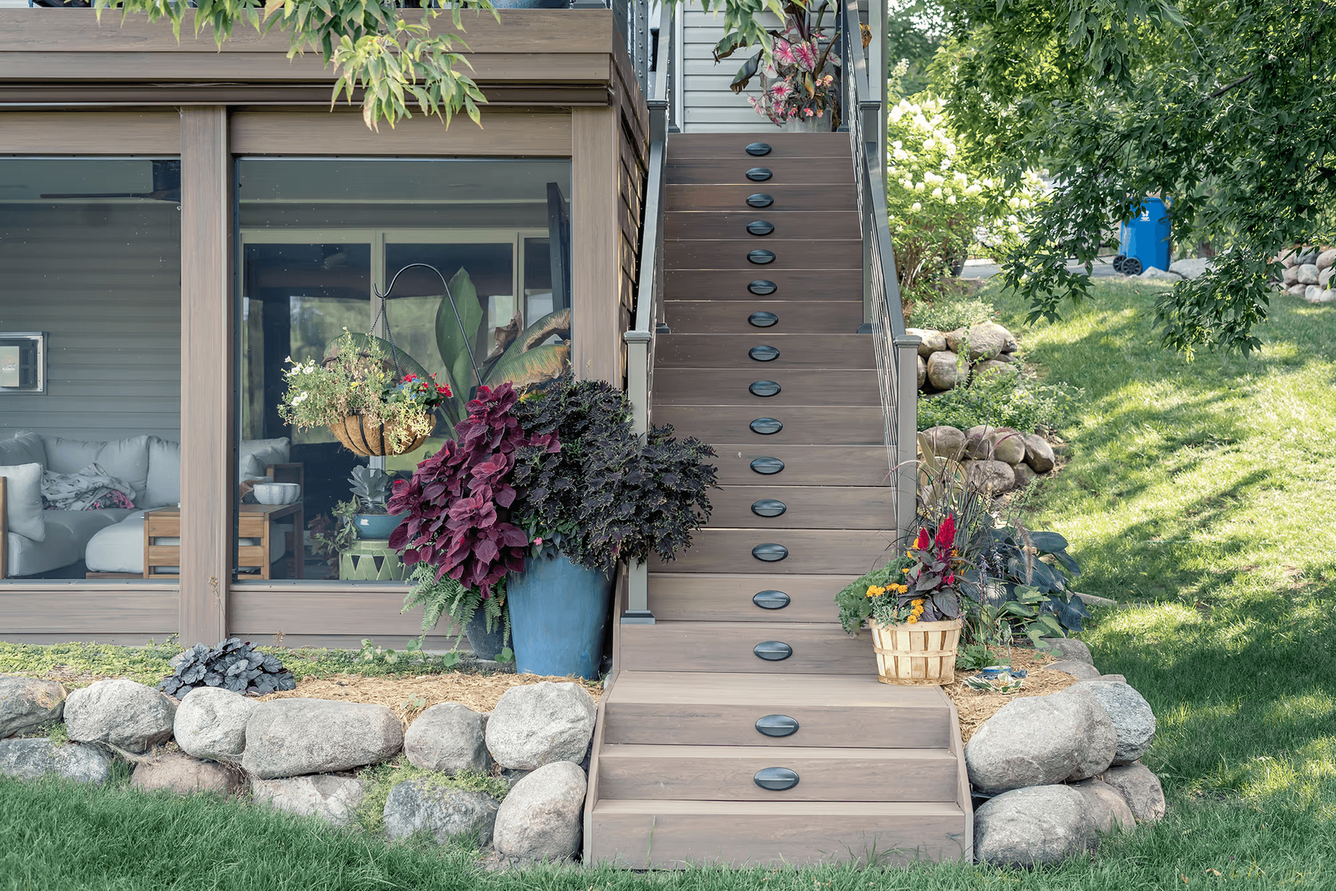 Ground-level view of composite deck stairs with embedded step lighting and cable railings leading to an upper level next to a glass-enclosed porch area with potted plants and rock landscaping.