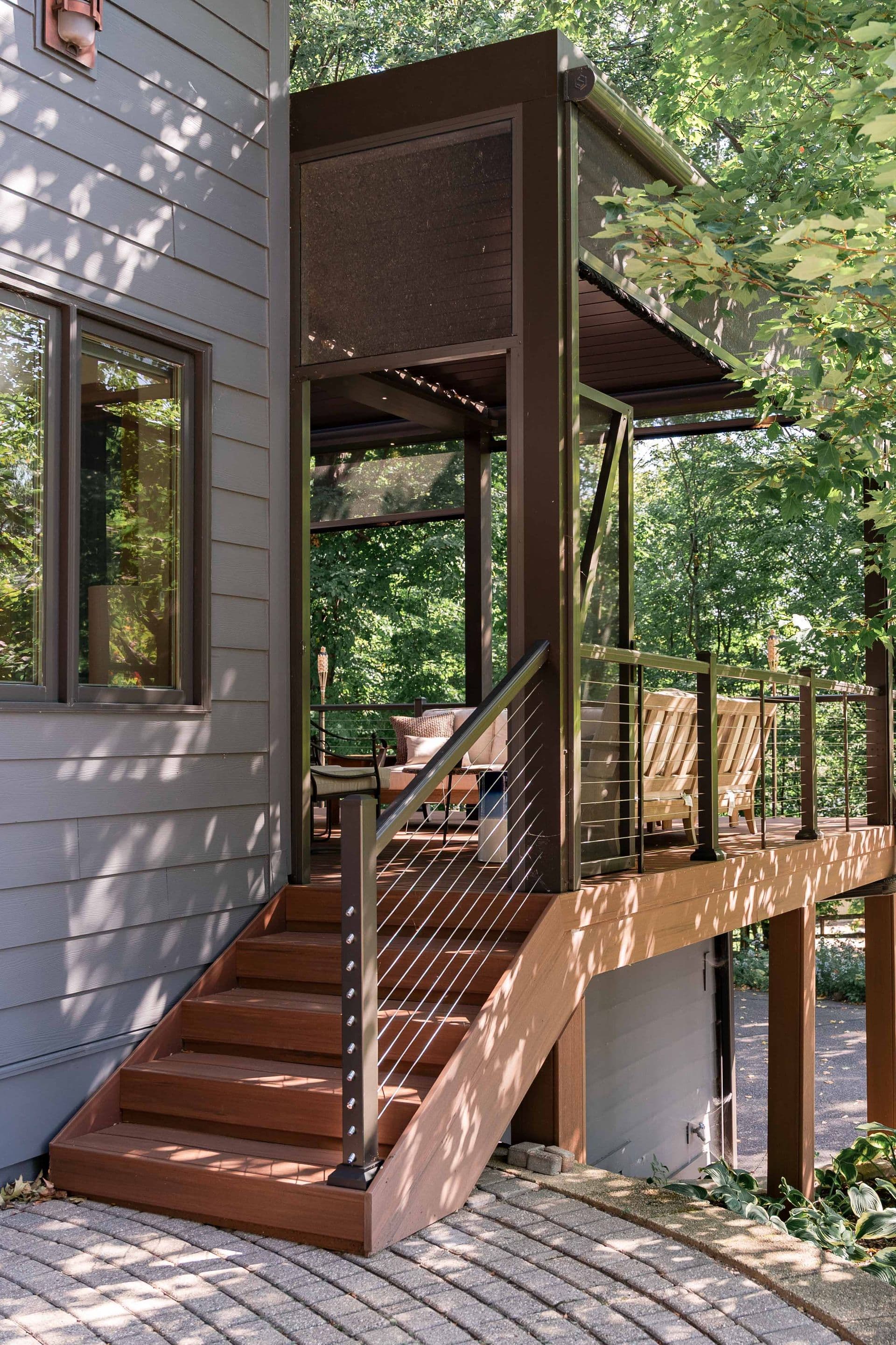 Eye-level view of composite deck stairs with dark cable railings leading up to a covered deck structure with patio seating attached to a grey siding house.