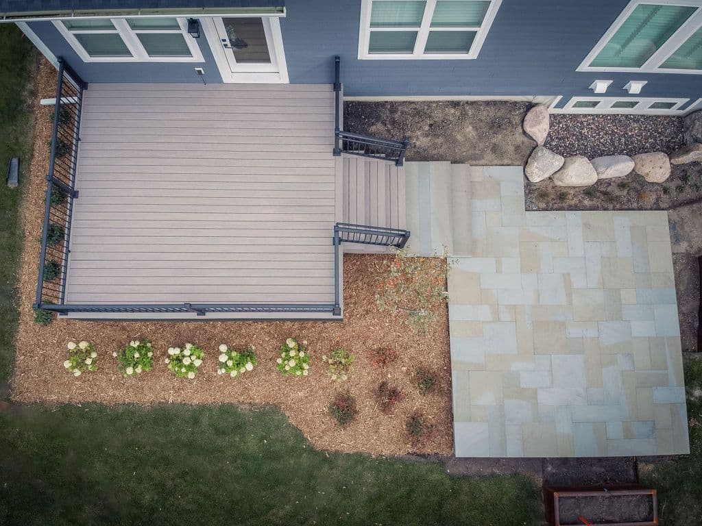 Direct overhead aerial view of a rectangular light grey composite deck attached to a blue home, featuring stairs down to an adjoining stone paver patio and surrounding mulch beds.